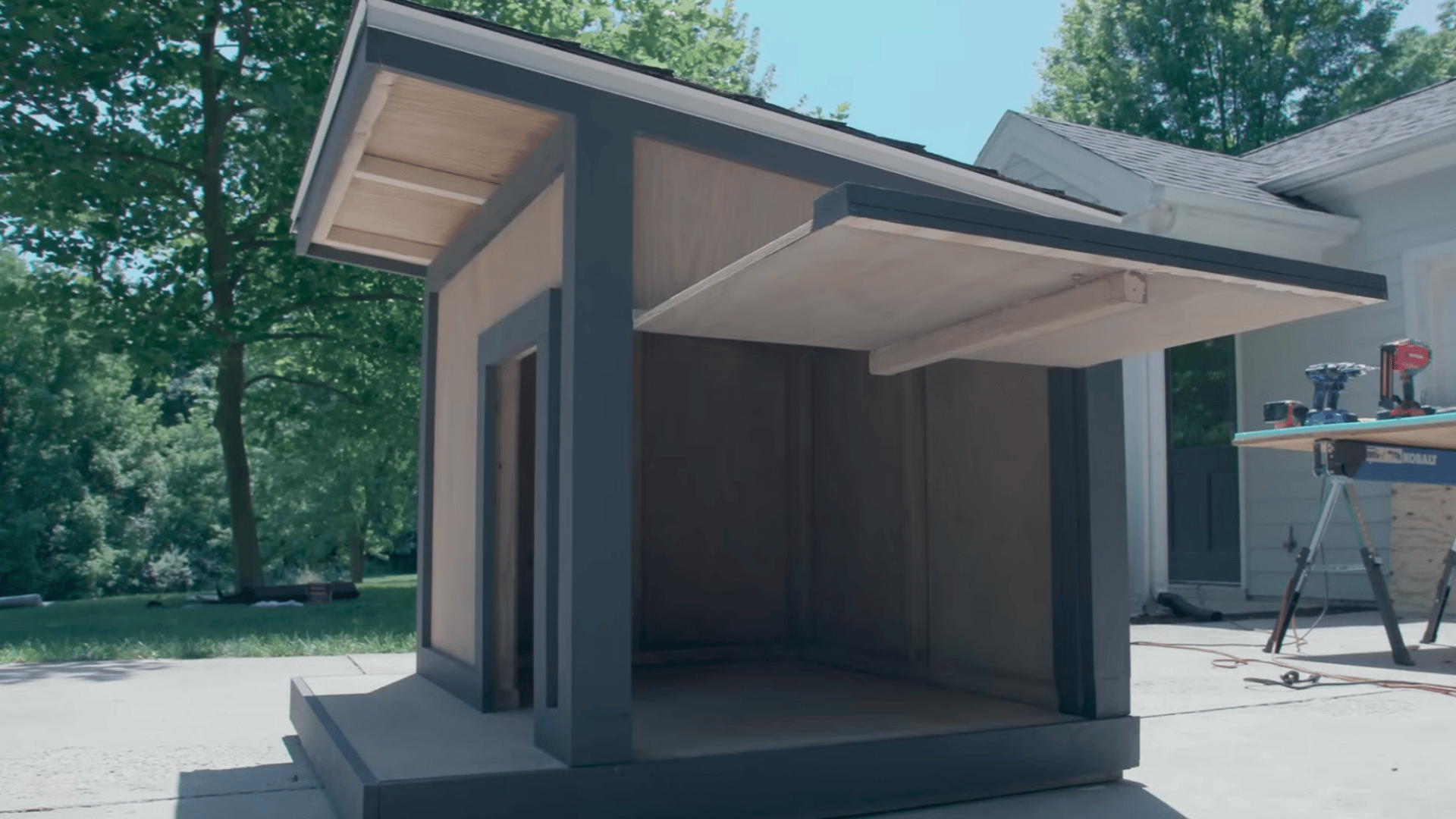 finished wooden pet shelter with a covered side opening and sloped roof displayed on a driveway beside a work table outdoors
