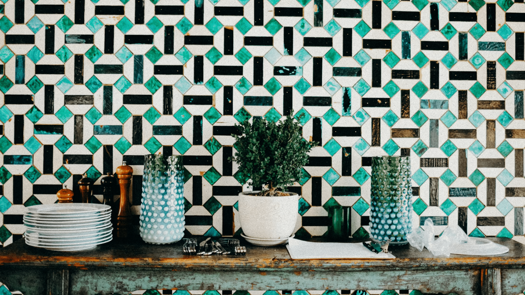 geometric turquoise black and white tiled wall behind a rustic table with glass vases stacked plates and a potted plant
