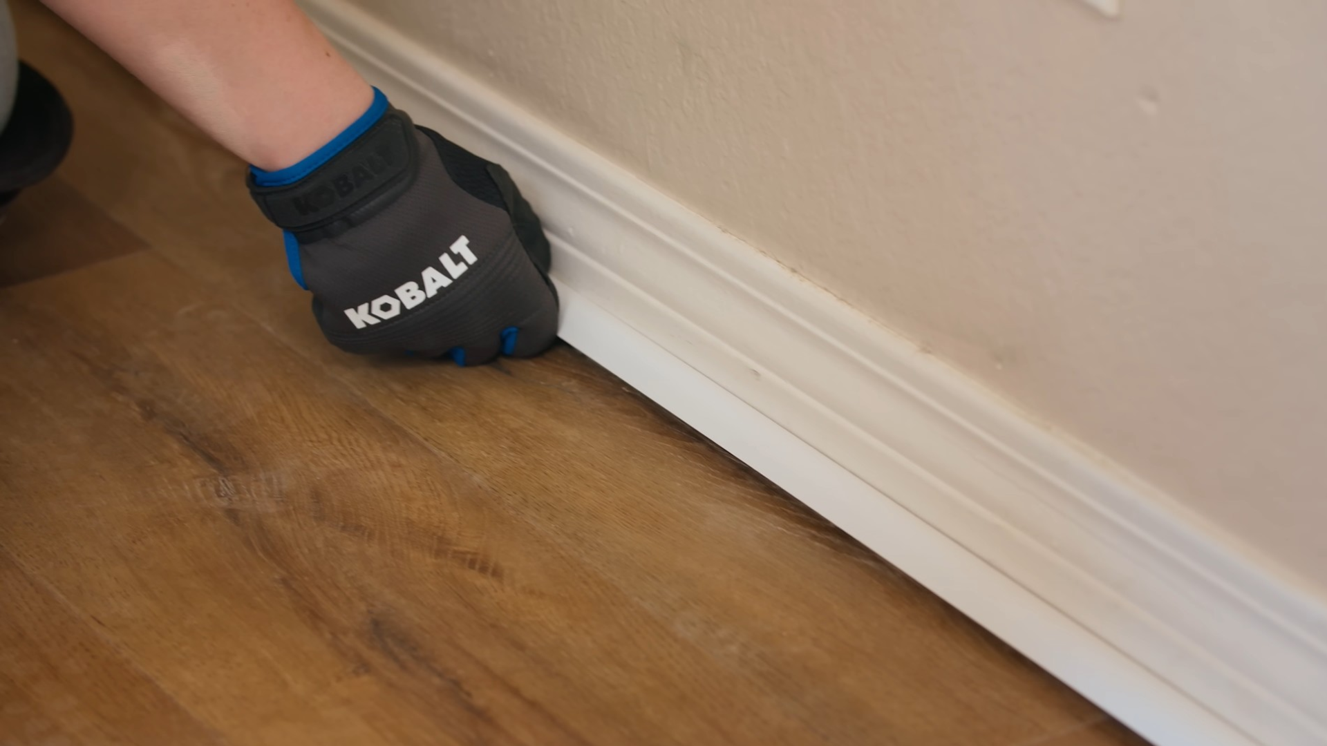gloved hand removes a spacer from the expansion gap between newly installed vinyl plank flooring and a white baseboard wall trim
