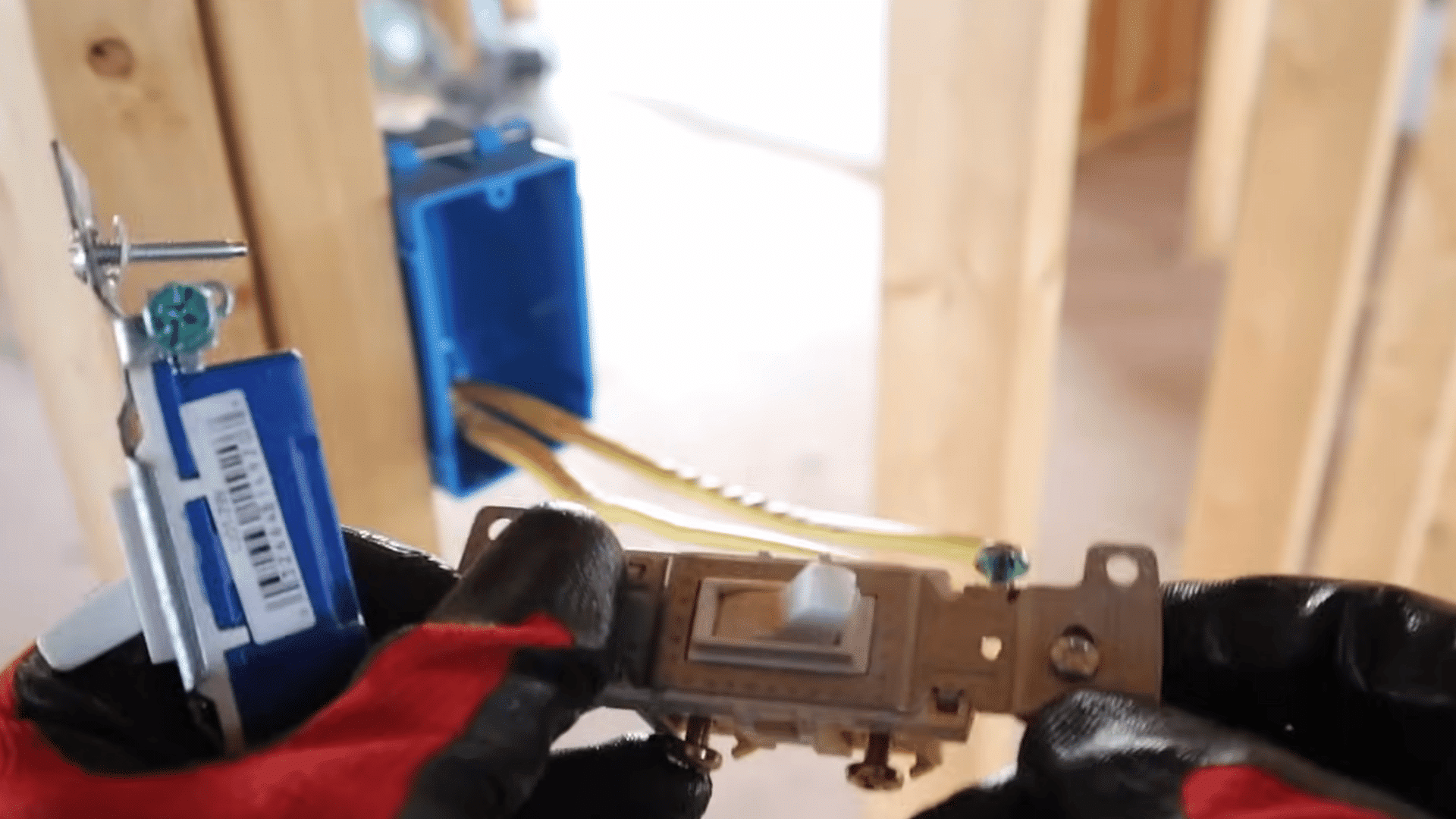 gloved hands comparing electrical light switches near a blue wall box with exposed wiring during installation work