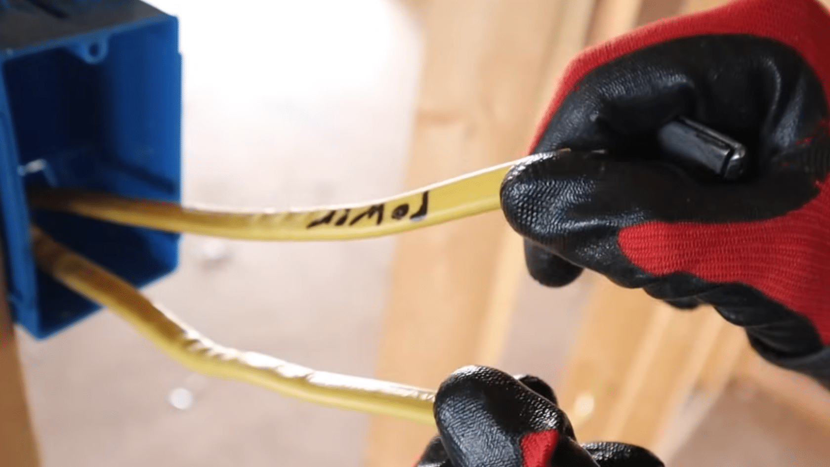 gloved hands hold labeled electrical wires extending from a blue wall box during a wiring installation project