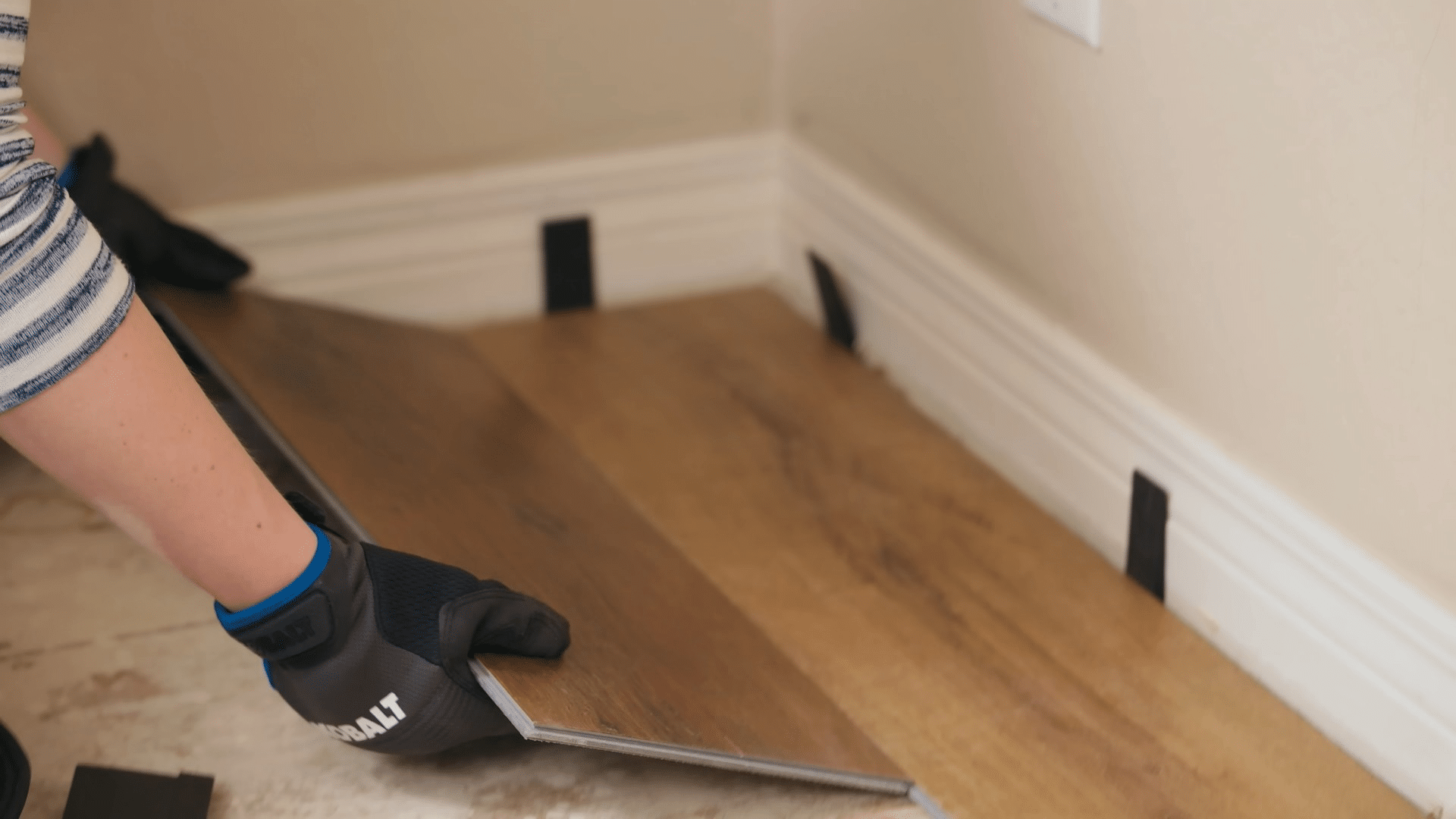 gloved hands position a wood look vinyl plank against spacers along a wall during flooring installation in a room corner