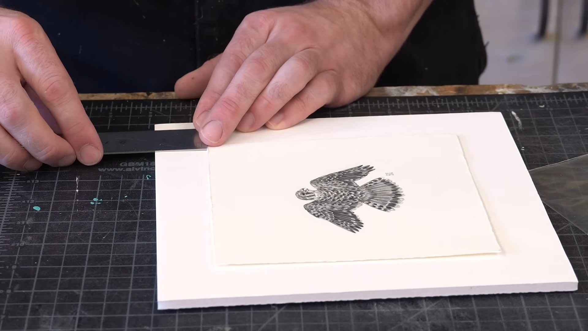 hands aligning a matted bird illustration with a metal ruler on a cutting mat during artwork framing preparation