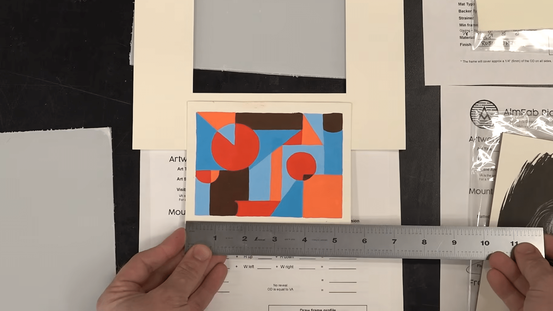 hands holding a metal ruler below a colorful geometric artwork sample placed on framing materials and mat board