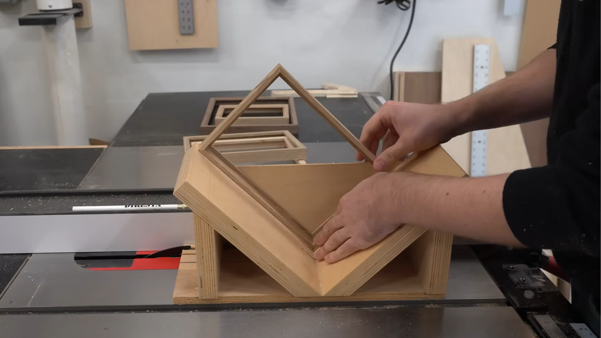 _hands positioning a wooden frame jig on a table saw in a workshop with stacked frames and tools in background