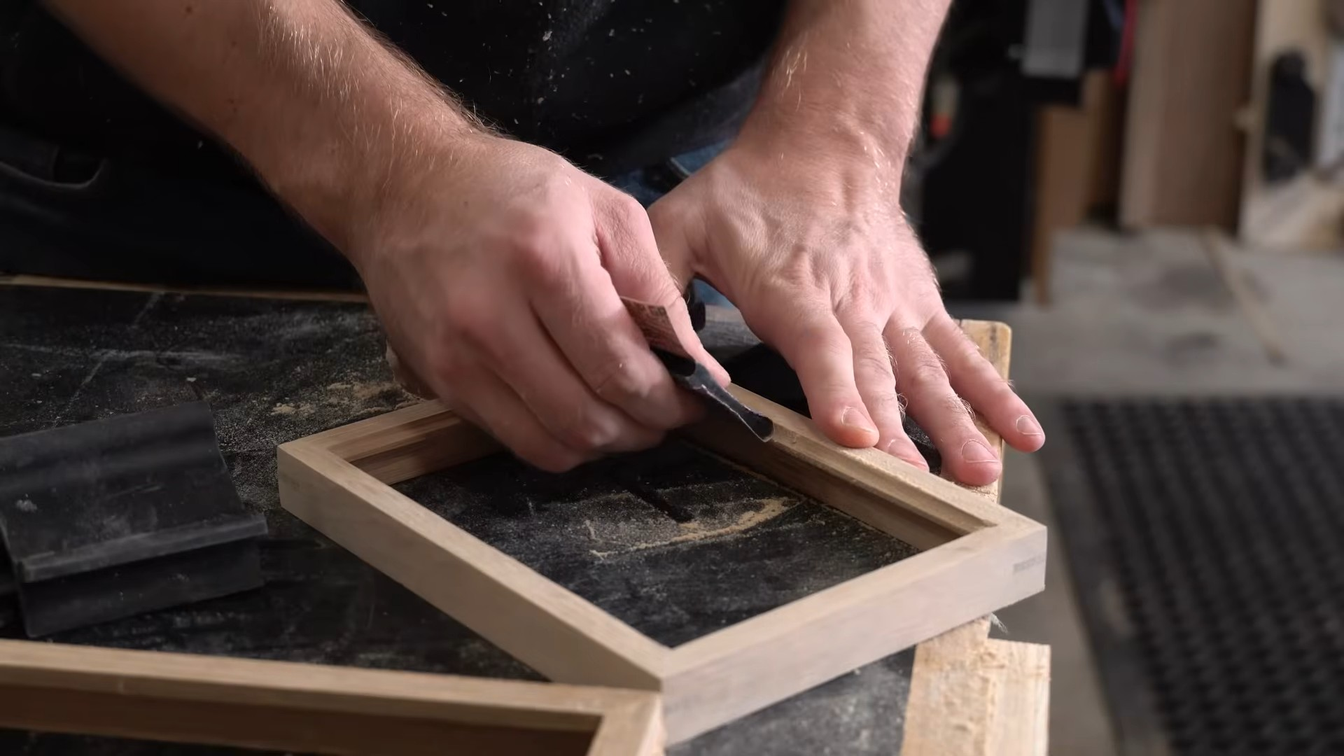 hands sanding the edge of a small wooden frame on a workbench with fine dust visible in a woodworking shop setting