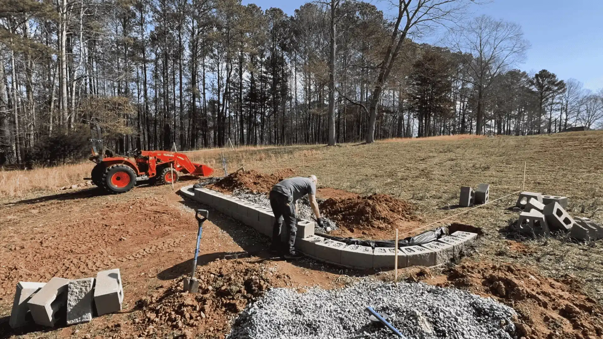 Worker building curved retaining wall with orange loader nearby, gravel drainage base in wooded construction site.