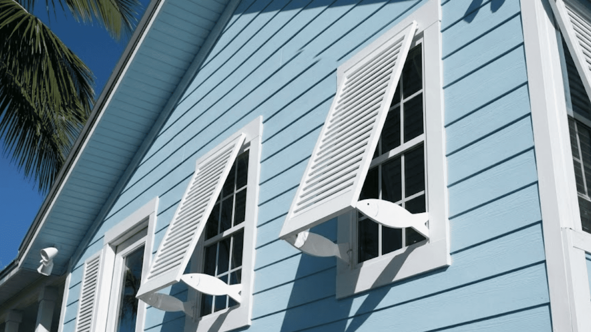 light blue house exterior with three windows flanked by open white louvers and a palm tree on the left