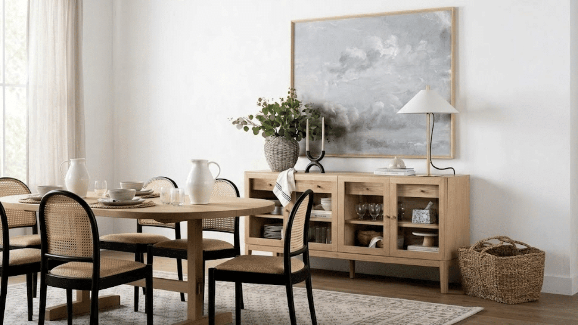 light wood dining table with woven chairs beside a matching sideboard decorated with greenery artwork and table lamp