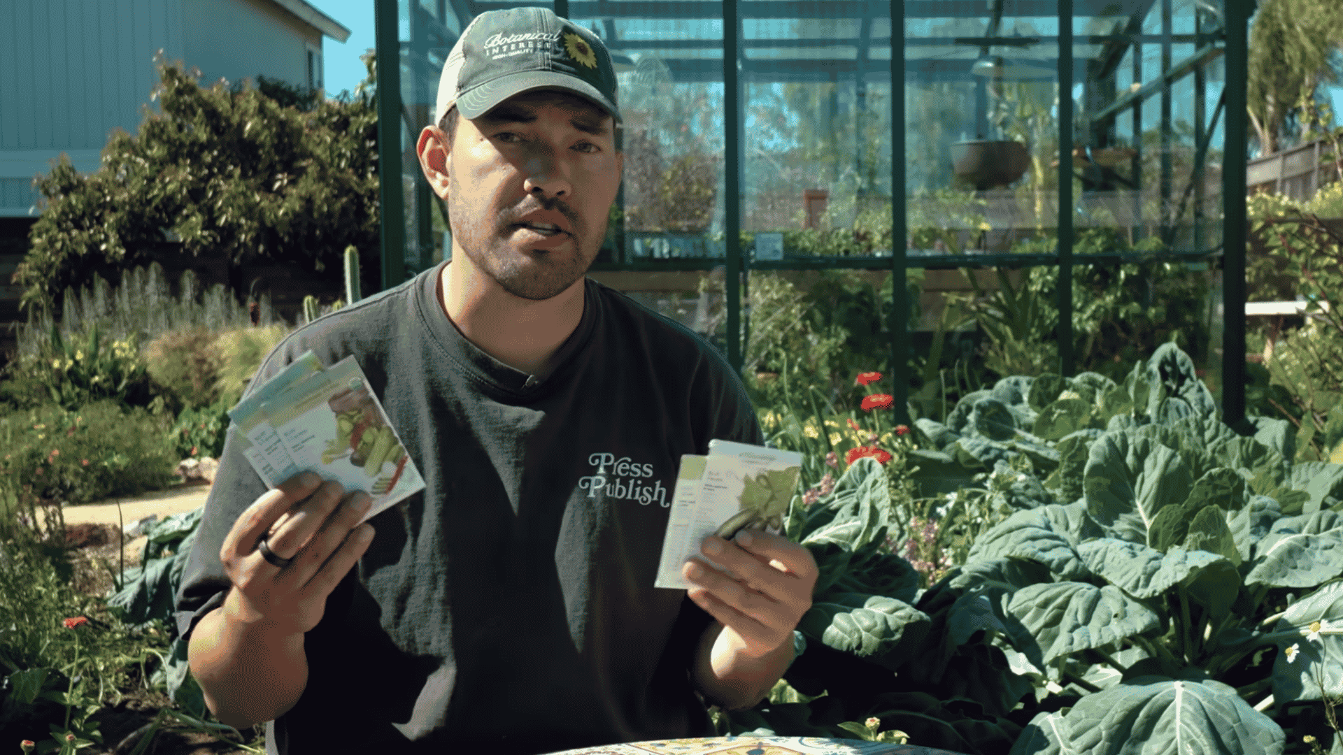 man holding seed packets in a vegetable garden with greenhouse and leafy plants growing in the background
