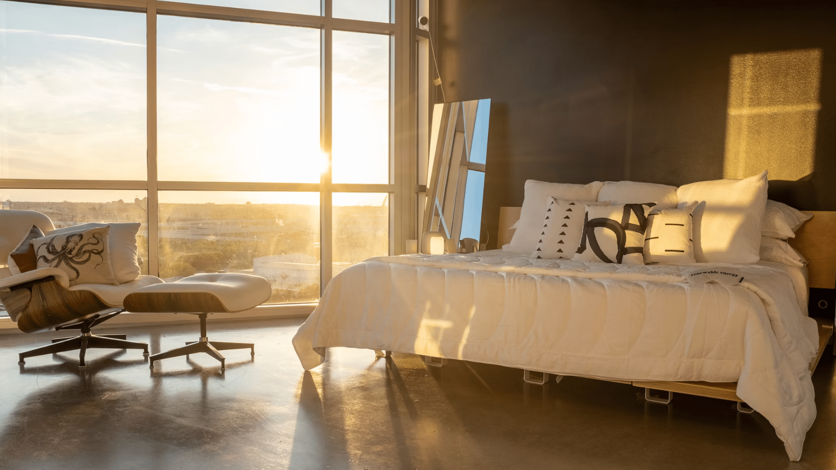 modern bedroom with a white bed beside floor to ceiling windows and lounge chair illuminated by warm sunset light indoors