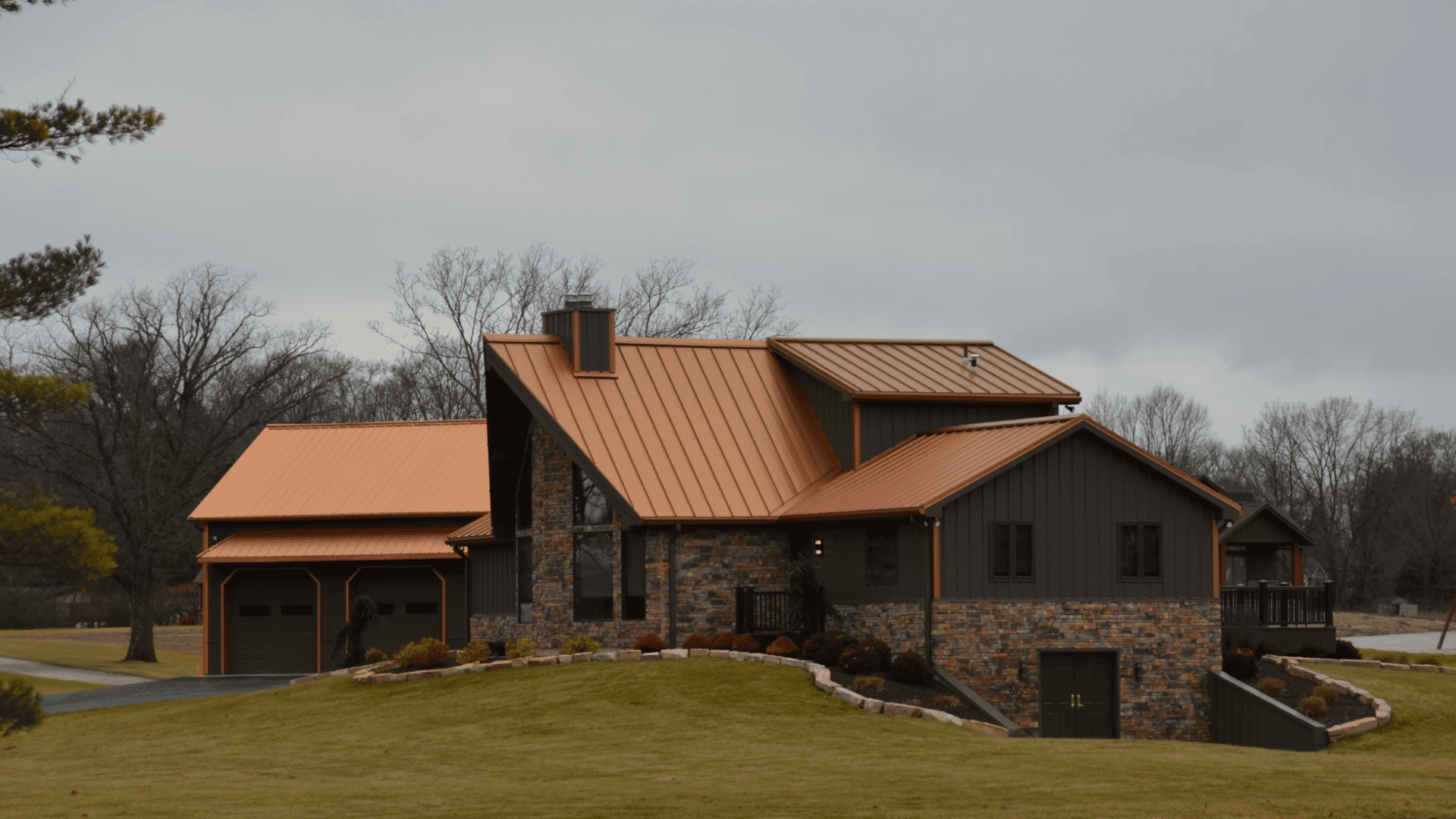 modern countryside home with copper metal roof stone exterior and landscaped lawn under gray skies