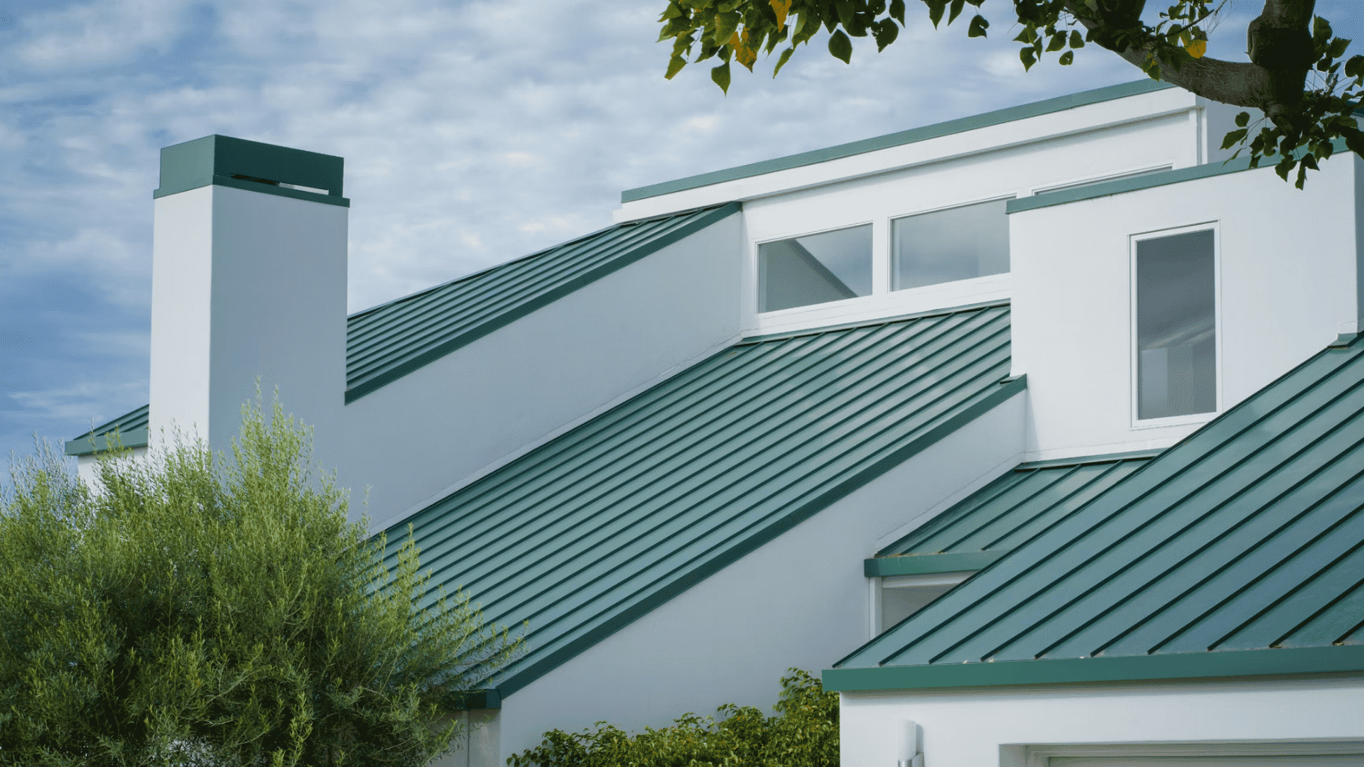 modern white house with green metal roof large windows and surrounding trees under a cloudy sky