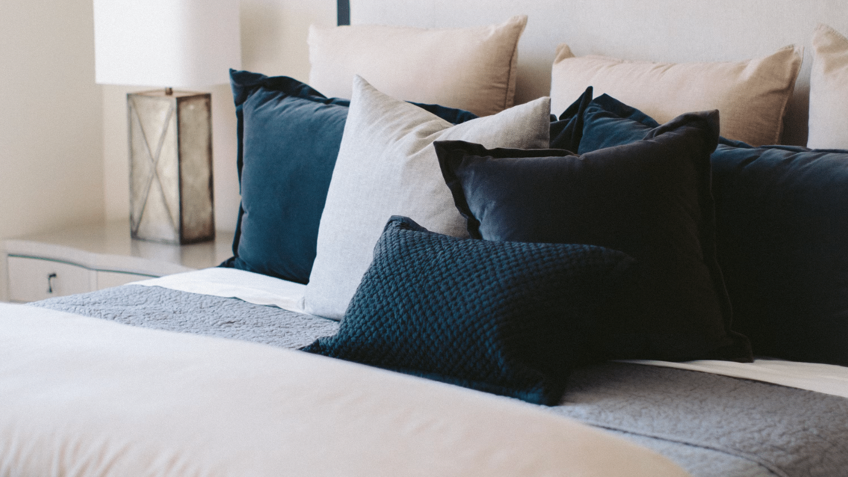 neatly made bed with layered blue and beige cushions soft bedding and a bedside lamp in a calm bedroom setting