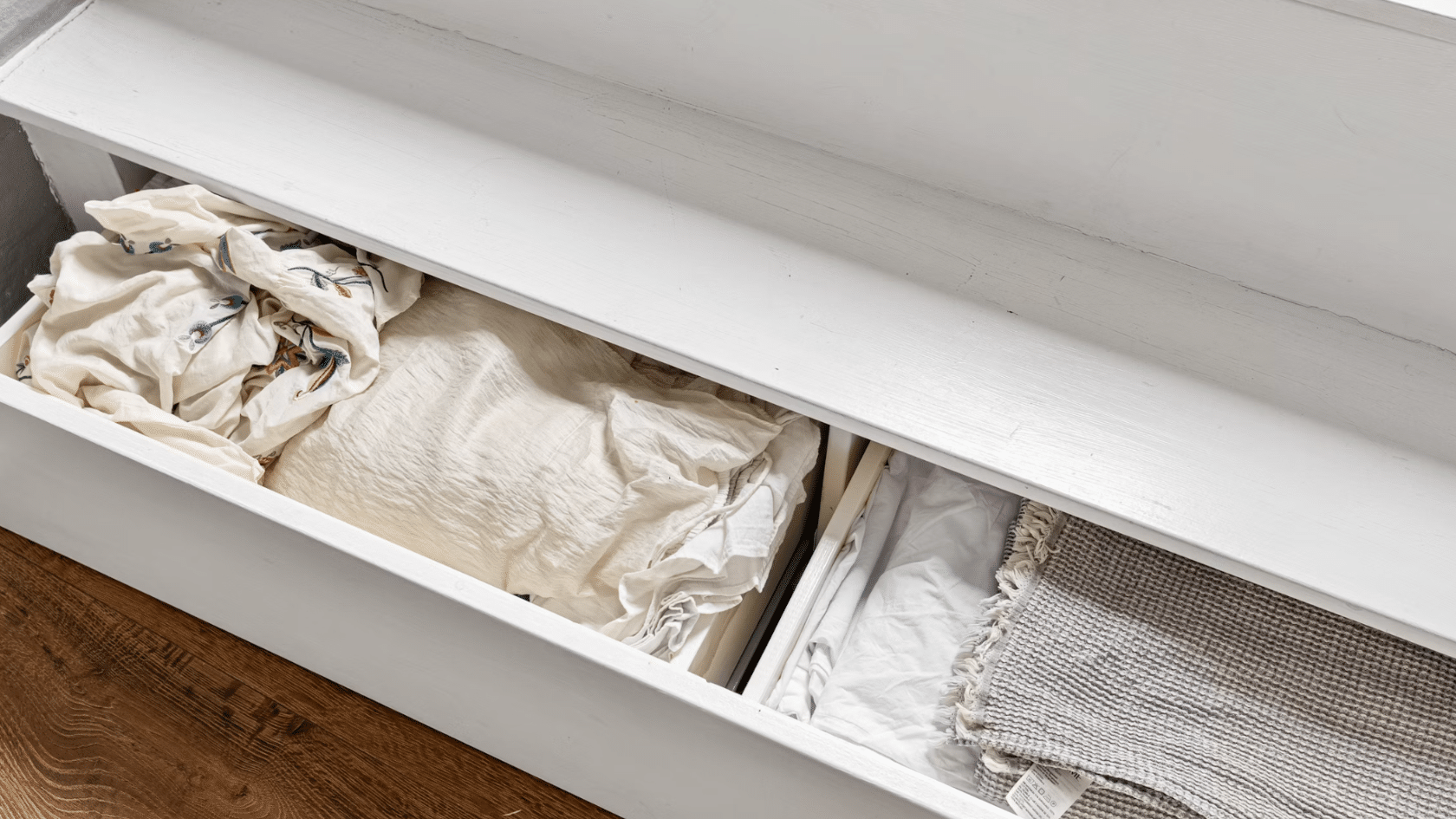open storage drawer beneath a white bench holding folded linens blankets and household textiles on a wooden floor