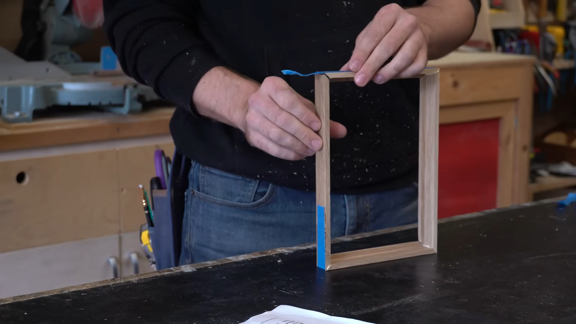 person aligning and taping together a small wooden frame with mitered corners on a workbench in a woodworking shop