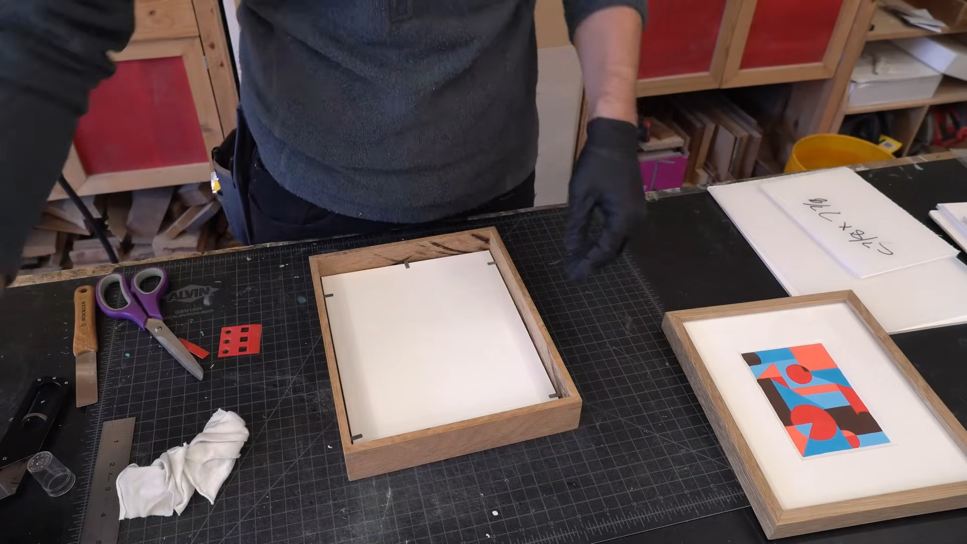 person assembling artwork inside a wooden picture frame on a cutting mat workbench with framing tools and supplies nearby