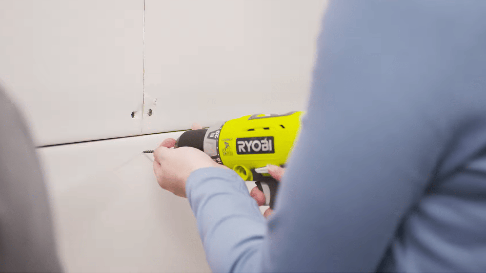 person driving a drywall screw into a wall panel with a power drill to secure the sheet to wall studs