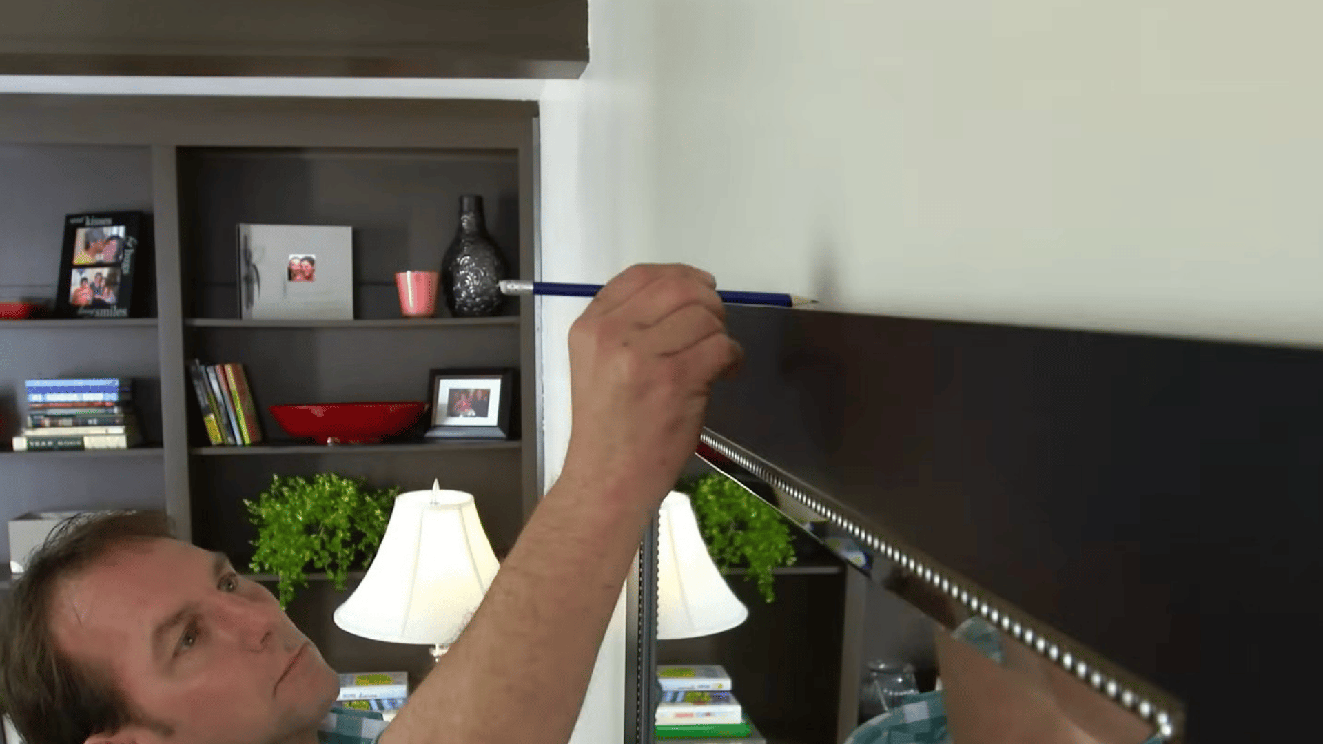 person holding a large mirror against a wall and marking the top edge with a pencil to position it for hanging accurately