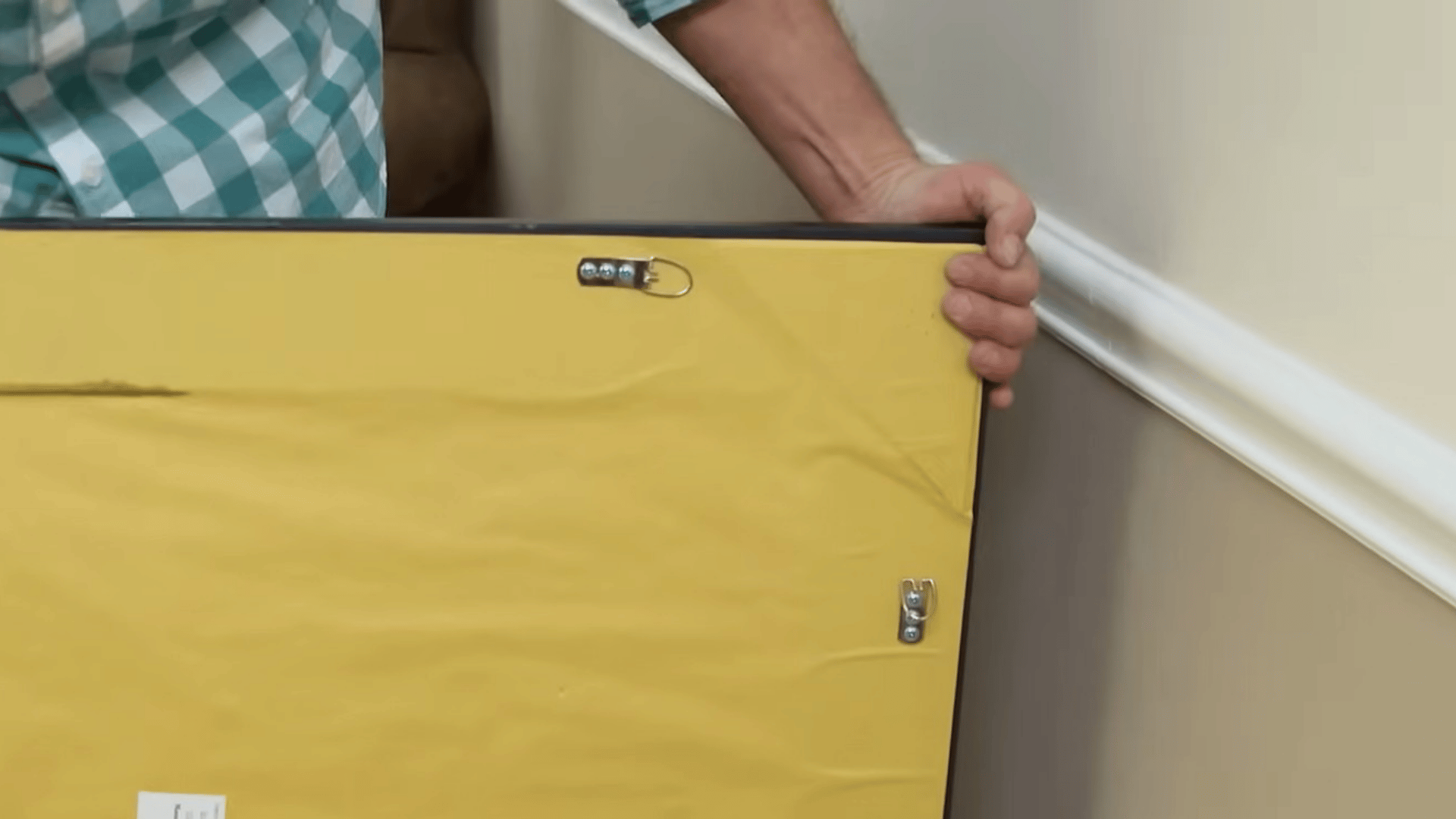 person holding the back of a framed picture showing hanging hardware while preparing to mount artwork on a wall