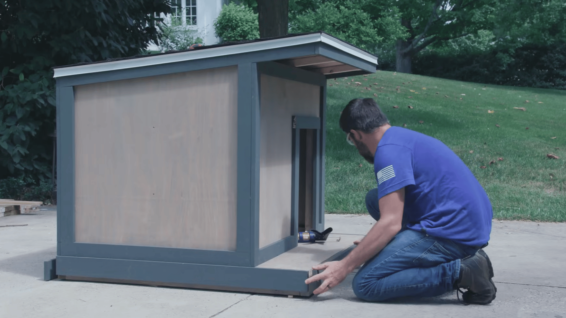 person kneeling beside a finished wooden pet shelter adjusting the base trim near the entrance on a driveway outdoors