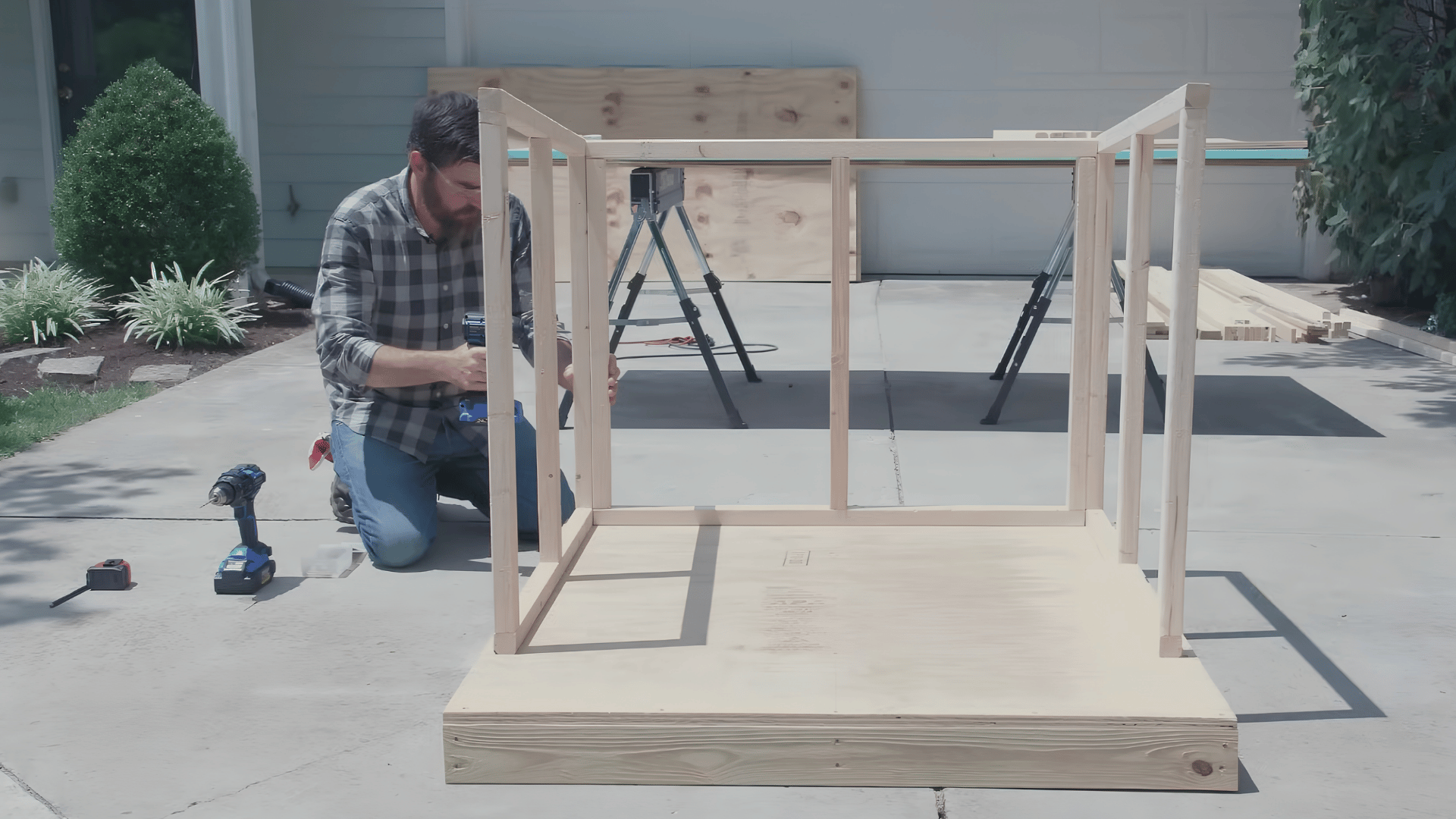 person kneeling beside a wooden platform using a power drill to assemble a framed structure on a driveway outdoors