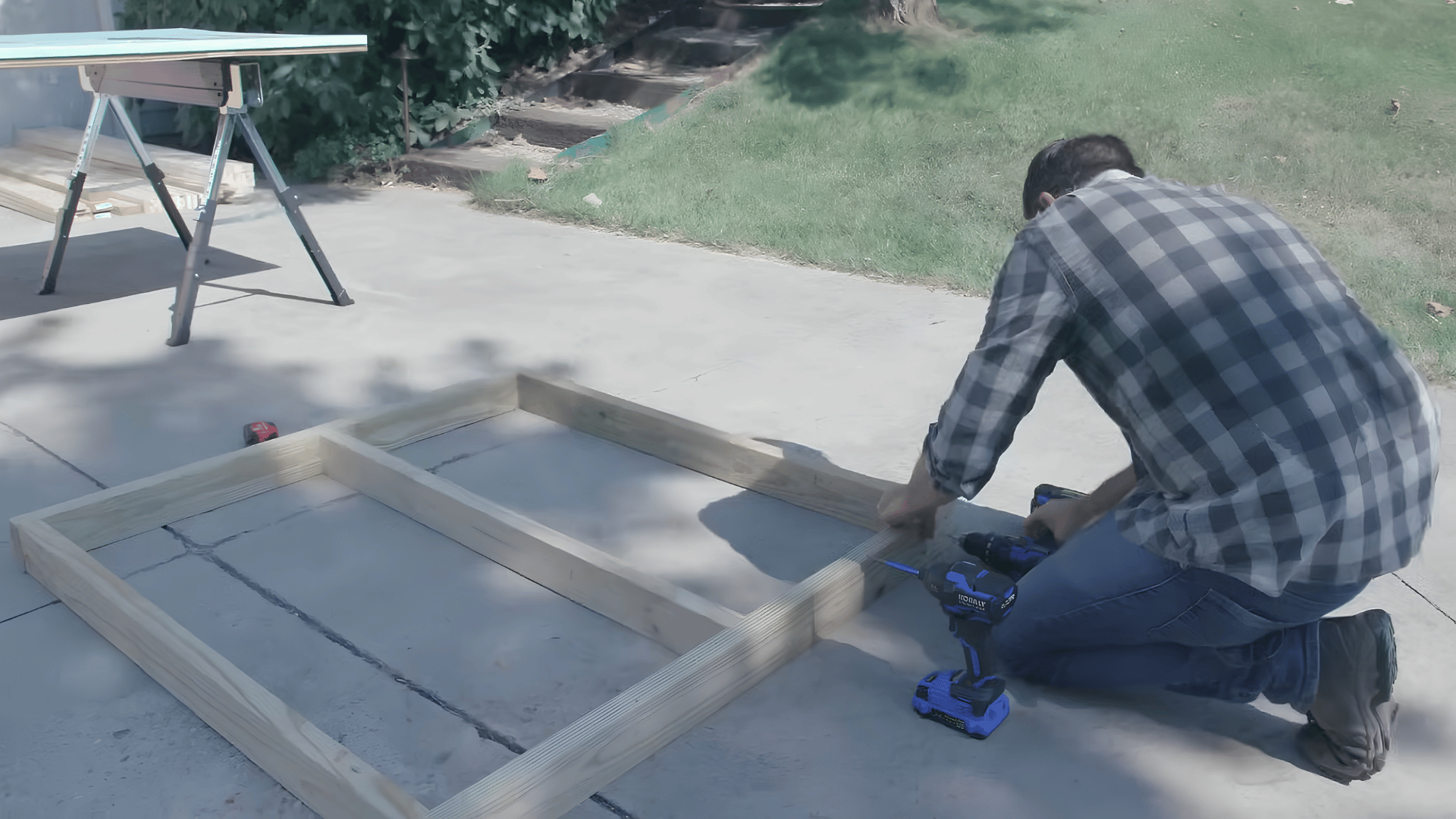 person kneeling on a driveway using a power drill to assemble a wooden frame beside a work table outdoors