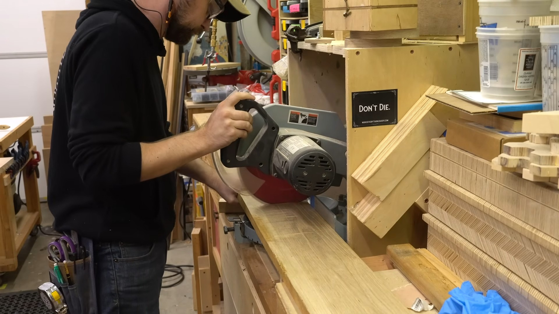 person operating a miter saw to cut a long wooden board in a woodworking shop surrounded by tools and lumber pieces