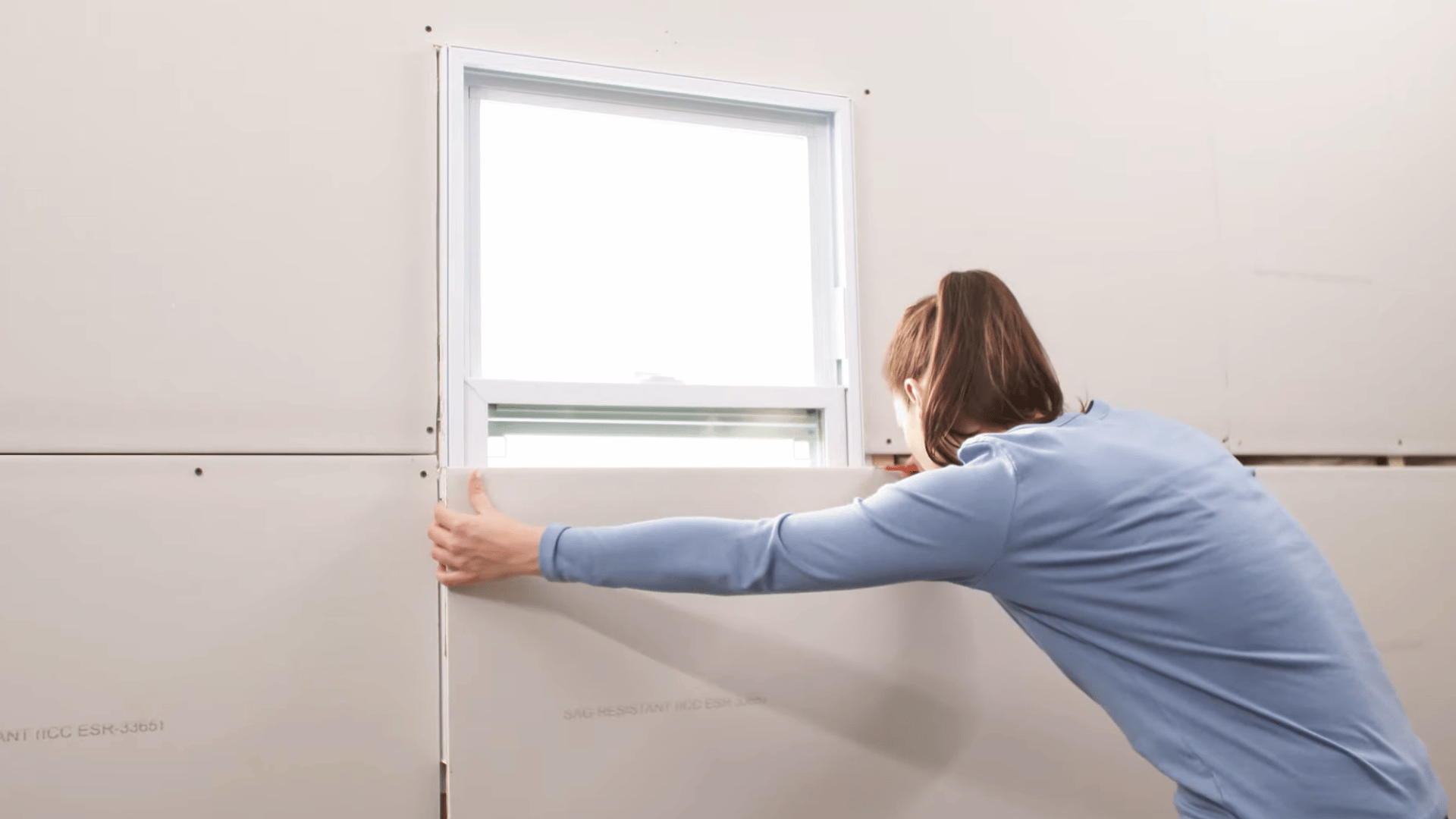 person positioning a drywall panel beneath a window opening while aligning edges for an accurate wall fit