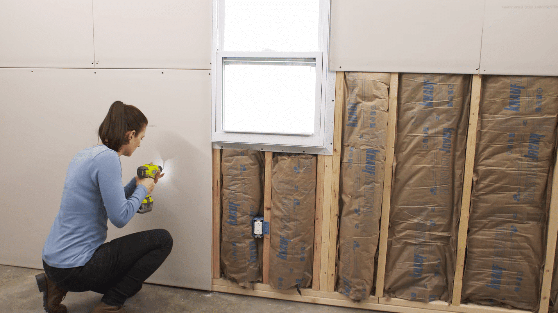 person securing a drywall panel beneath a window opening using a power drill during wall installation work