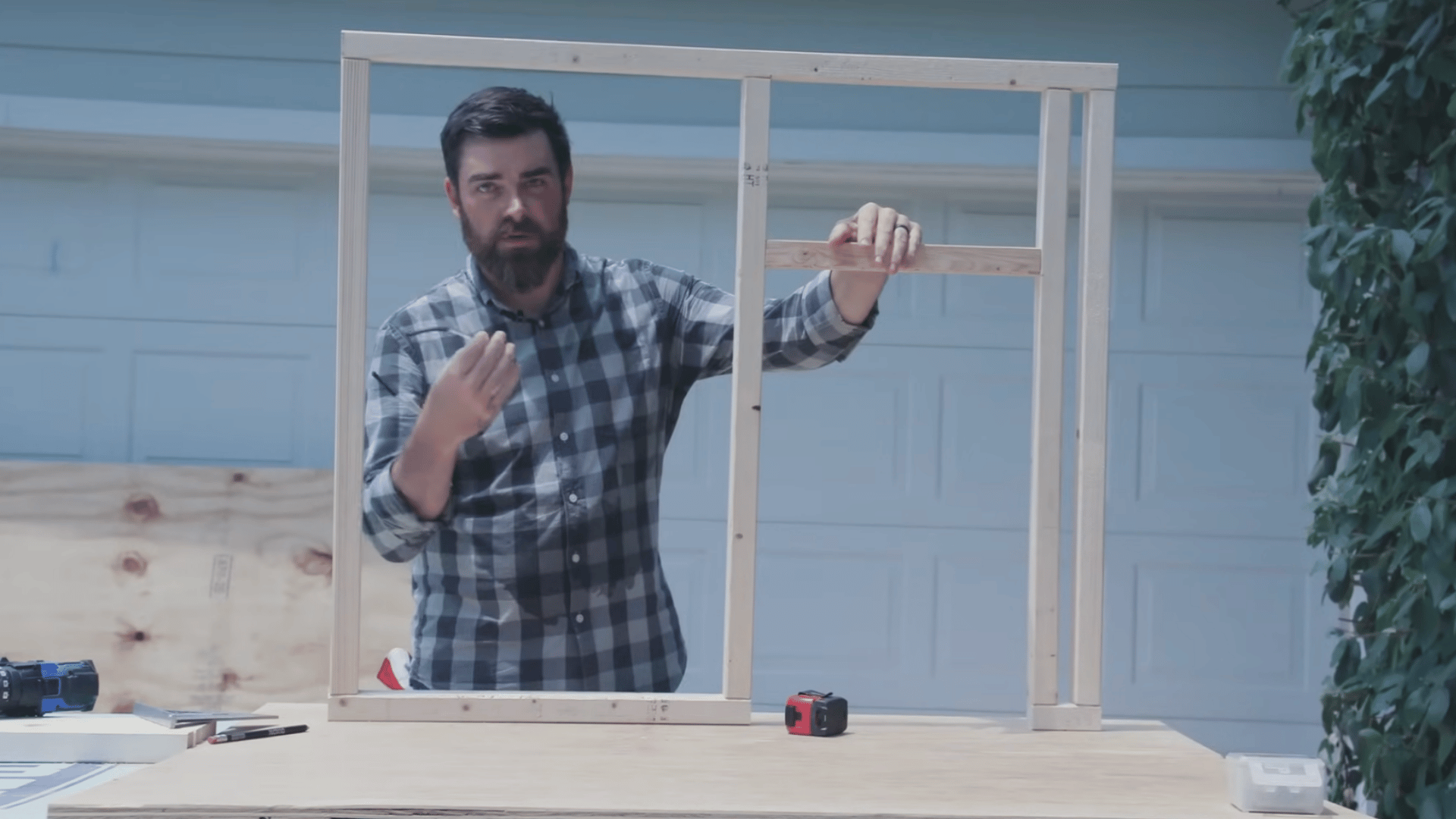 person standing behind a wooden wall frame on a workbench gesturing while explaining a construction project outdoors
