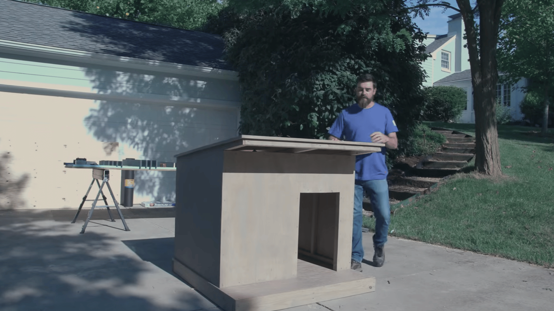 person standing beside a completed wooden pet shelter with a slanted roof and doorway on a driveway outdoors
