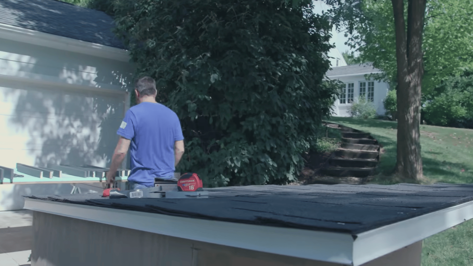 person standing beside a finished wooden pet shelter with a shingled sloped roof while working in a driveway outdoors