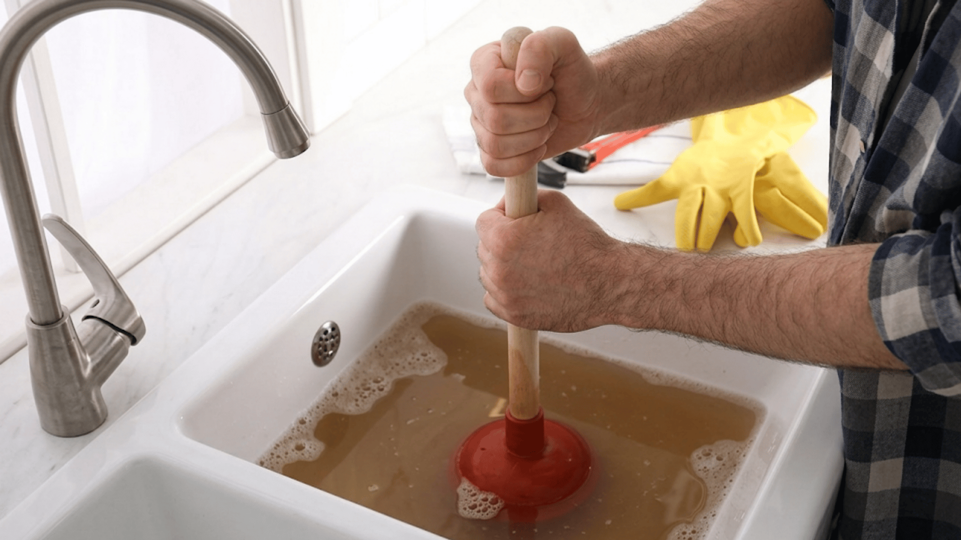person using a plunger to clear a clogged kitchen sink filled with dirty water and soap bubbles