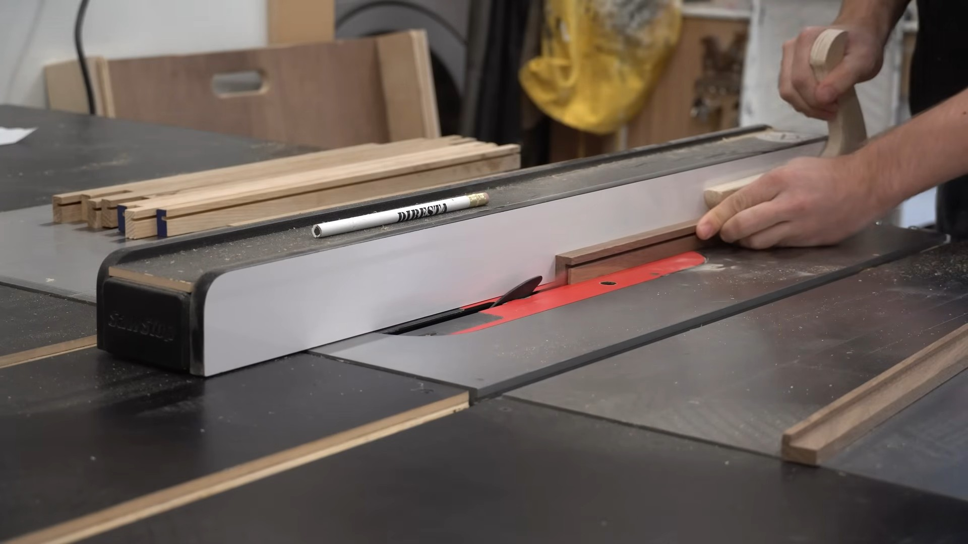 person using a push stick to guide a narrow wood strip through a table saw in a woodworking shop with lumber nearby
