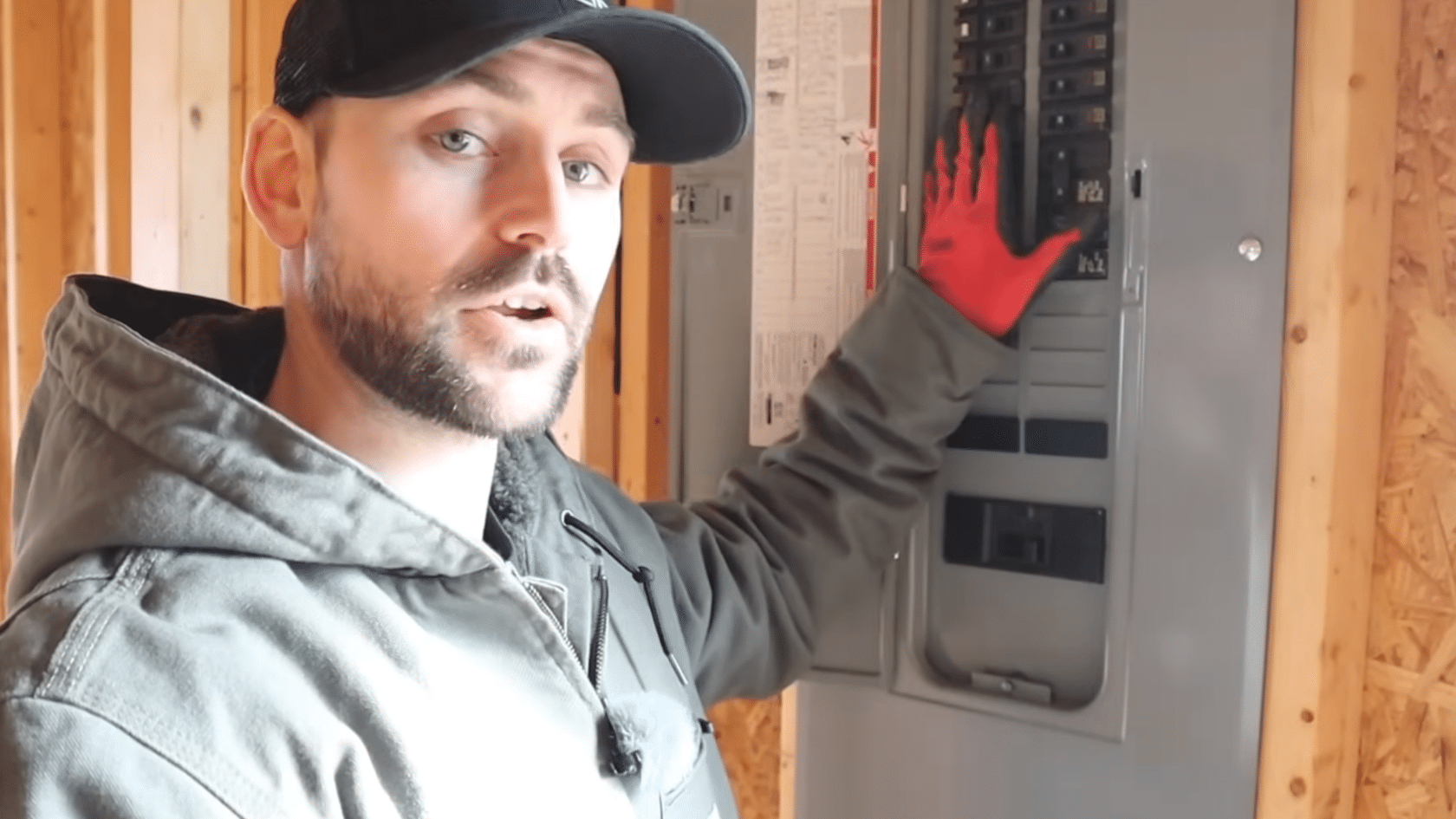 person wearing a cap and work jacket stands beside an electrical panel while demonstrating electrical work indoors