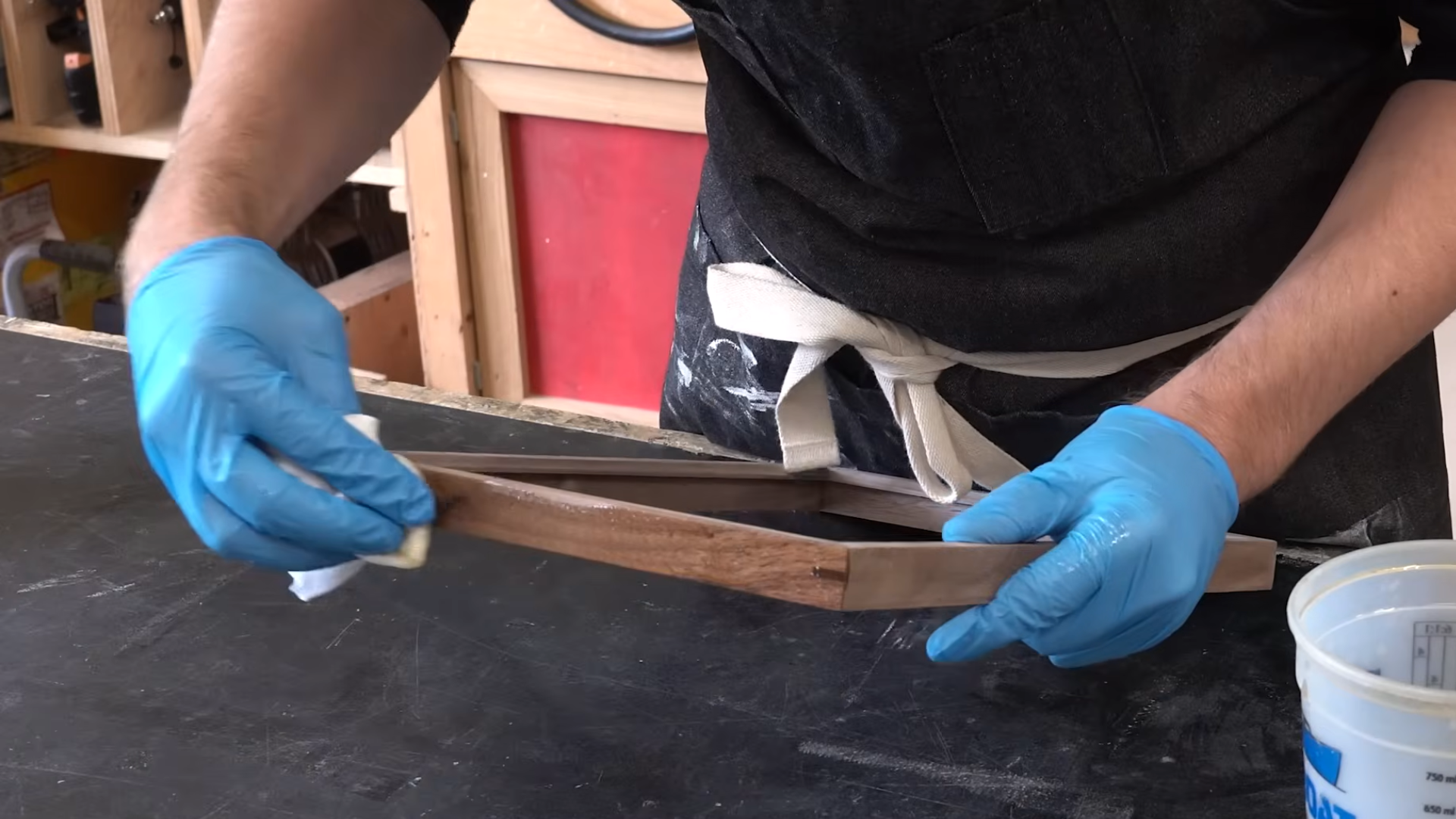 person wearing blue gloves applying finish to a wooden frame on a workbench with a cloth in a woodworking shop setting
