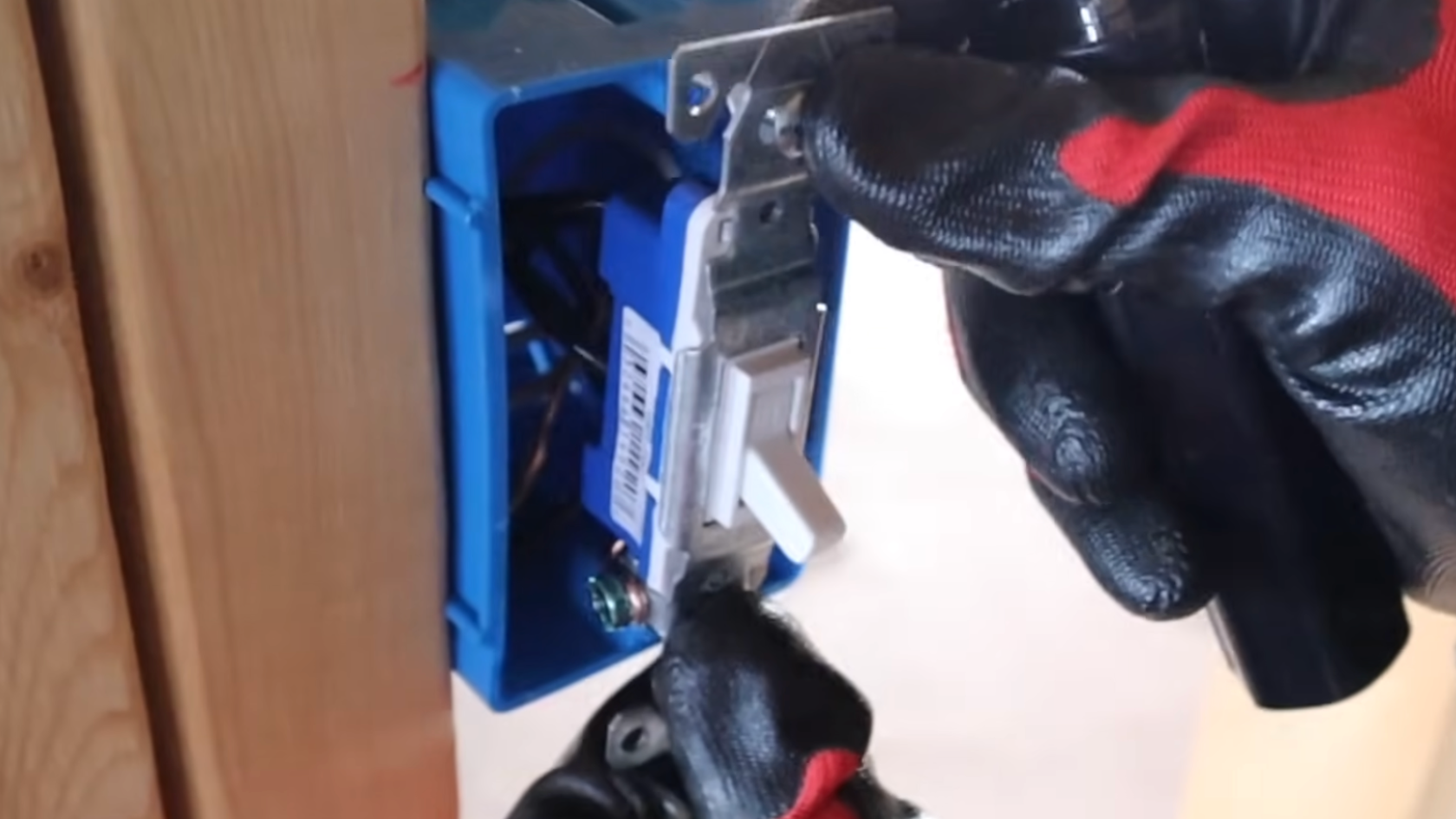 person wearing red and black work gloves installs a wall switch into a blue electrical box during wiring work indoors