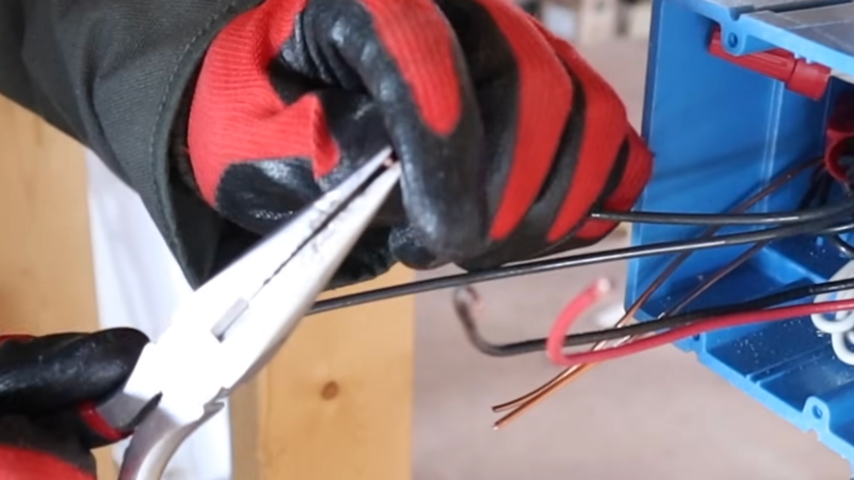 person wearing red and black work gloves uses needle nose pliers to bend electrical wires inside a blue junction box during installation