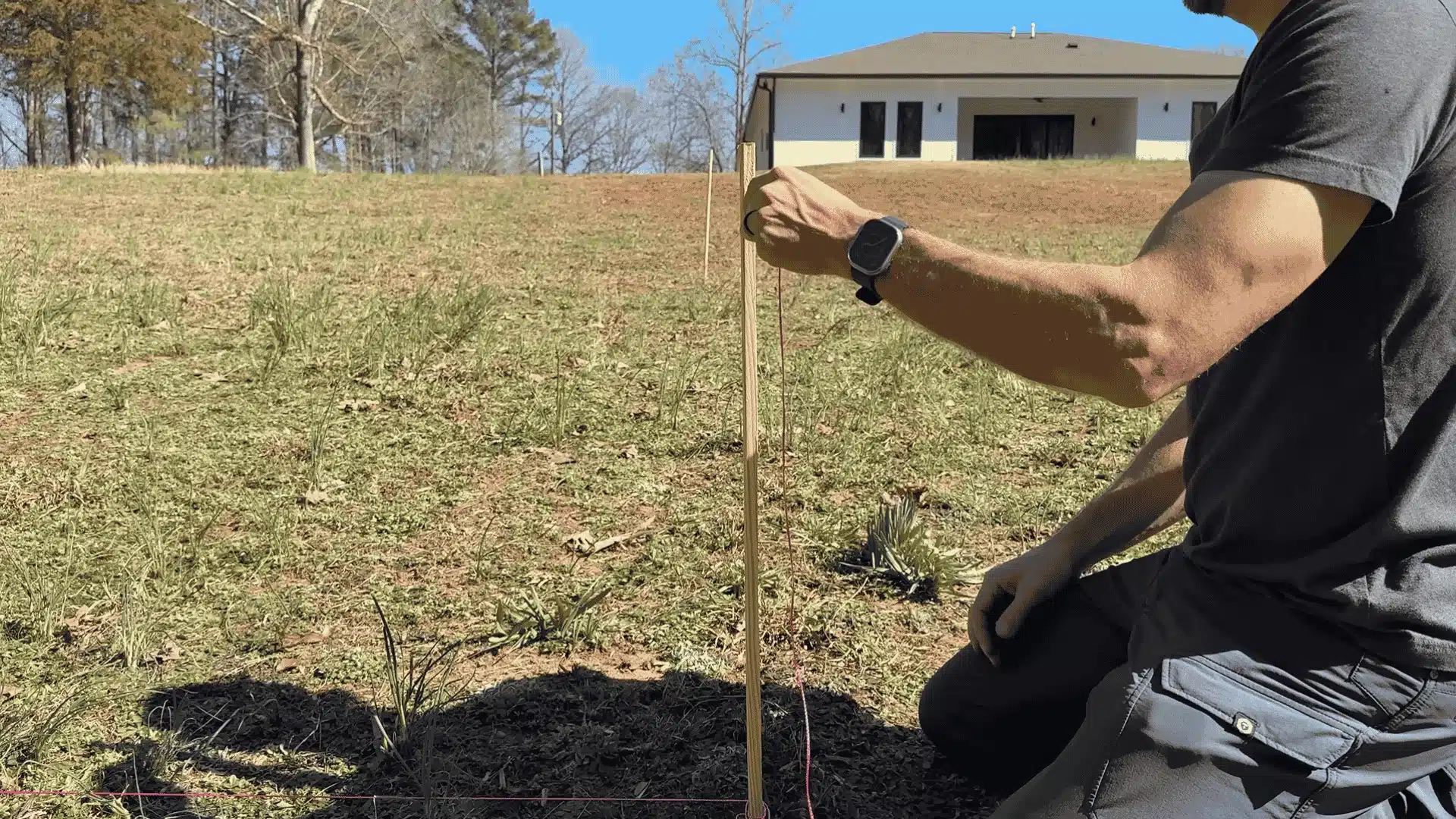 Person kneeling in a yard using a stake and string line to mark layout points in front of a house under construction.