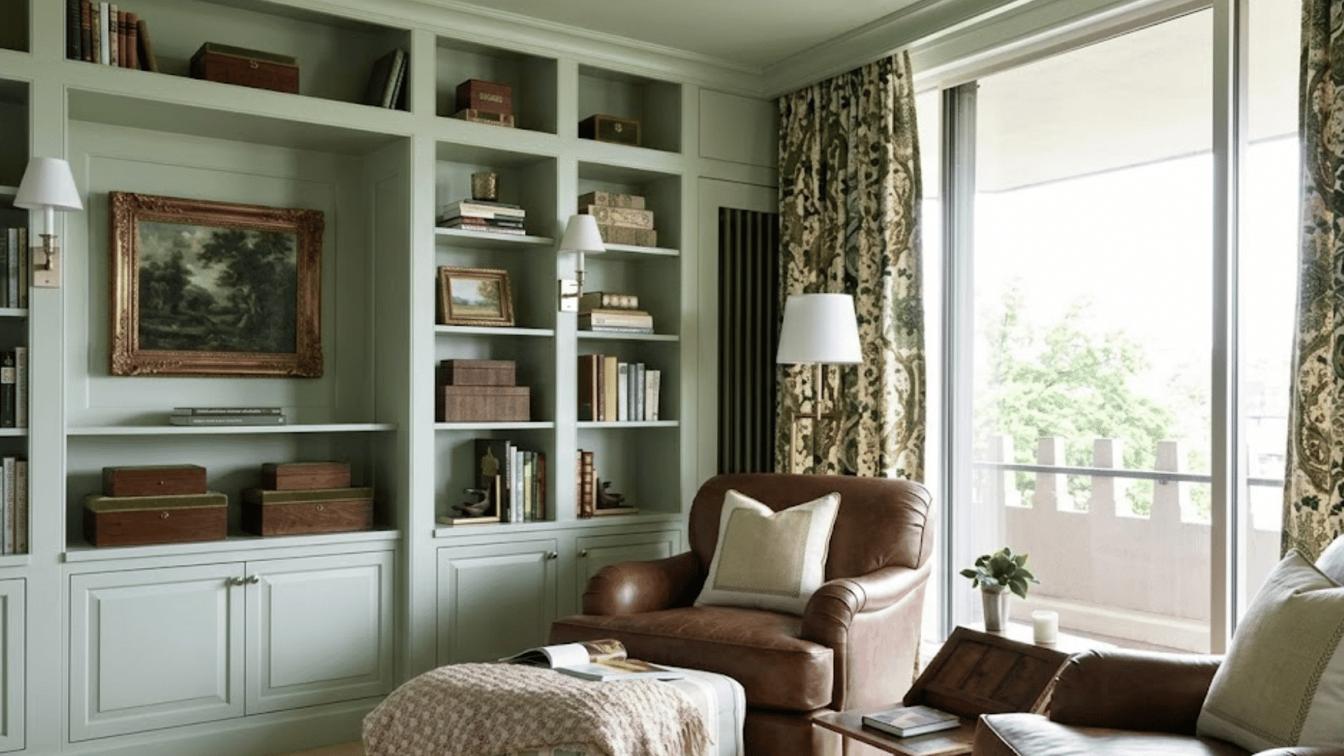 sage green built in bookshelves with two leather chairs and an ottoman by a window with floral curtains