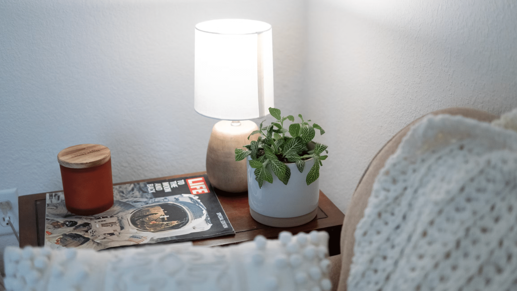 side table with a lit lamp potted plant candle and magazine beside a cozy chair with textured cushions and blanket