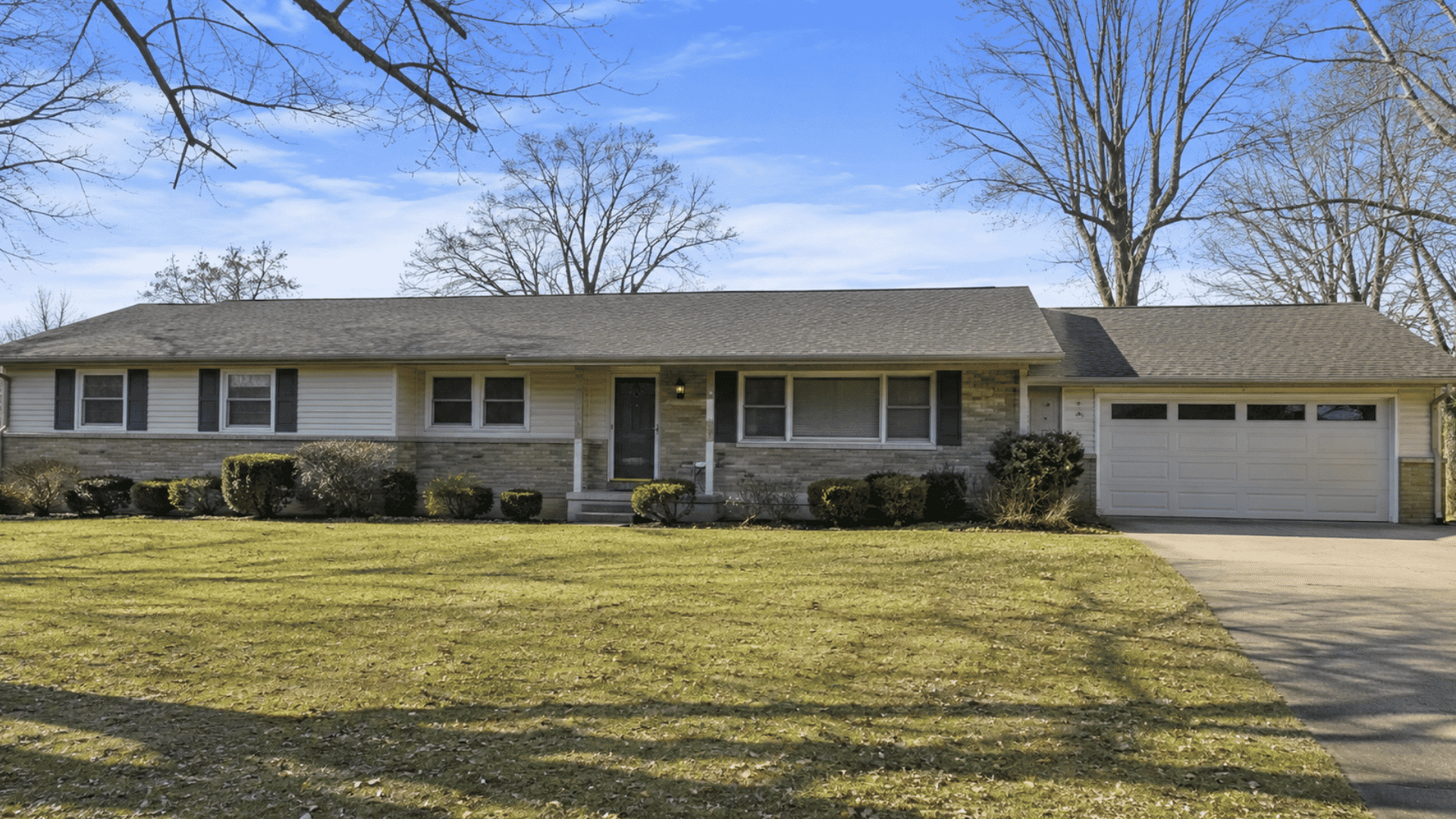 single story suburban house with attached two car garage trimmed shrubs and broad lawn beneath bare trees