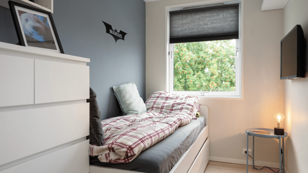 small bedroom with a single bed plaid bedding and window view alongside a dresser wall mounted television and side table lamp