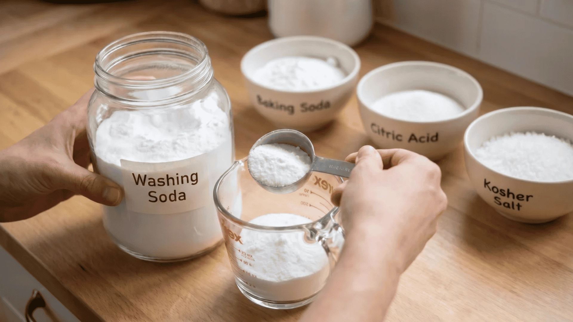 Hands measuring washing soda into a cup with bowls of baking soda, citric acid, and kosher salt visible in the background