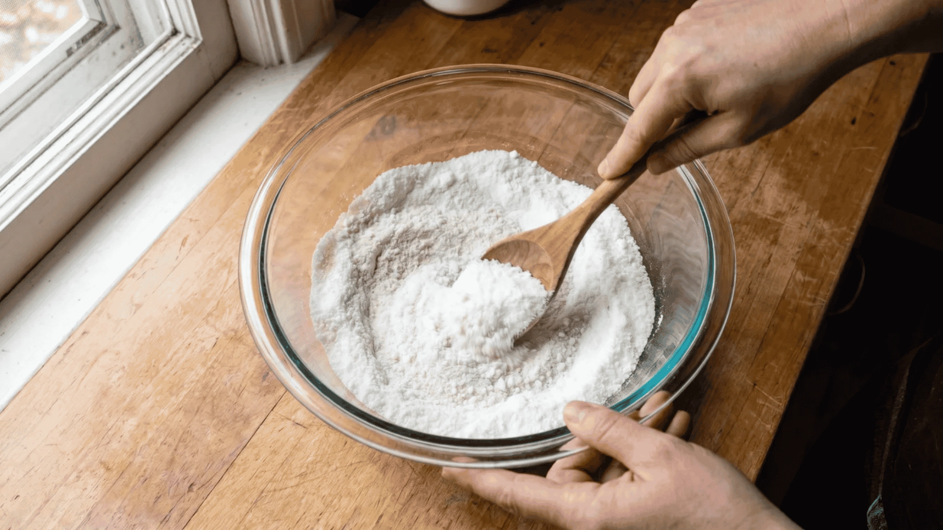 Hands stirring a large glass bowl containing washing soda, baking soda, citric acid, and salt until the mixture is uniform and clump-free.