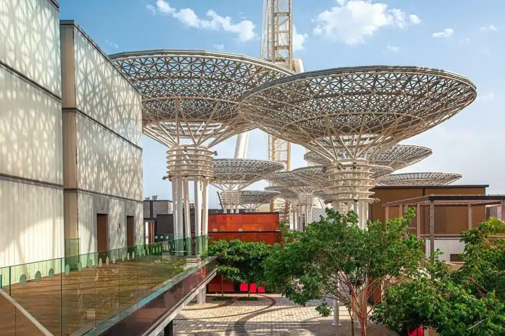 Futuristic architectural structures with lattice canopies over walkway and trees in an outdoor urban setting