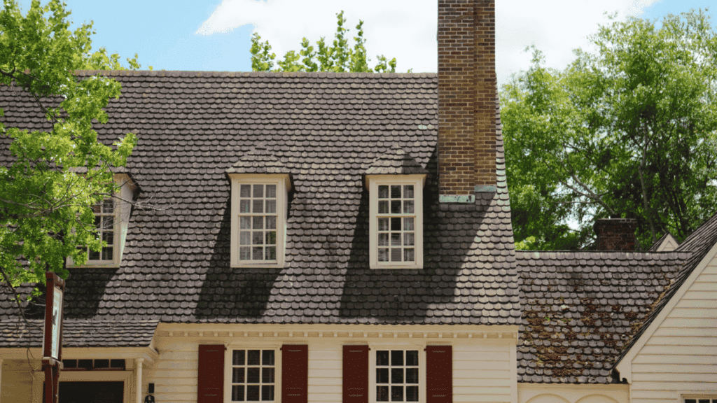traditional house with dark shingle roof dormer windows and brick chimney surrounded by green trees