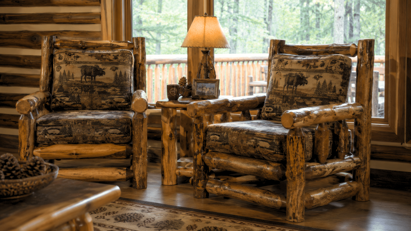 two rustic log armchairs with wildlife patterned cushions beside a wooden table and lamp near large forest view windows