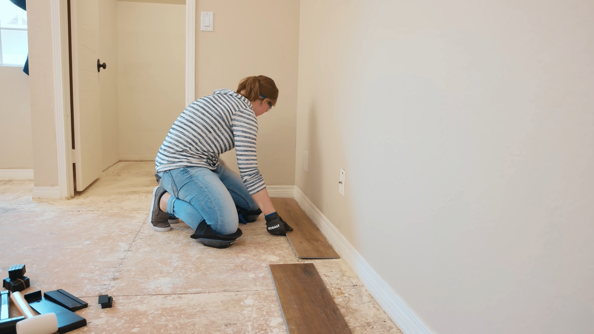 woman kneels on an exposed subfloor positioning vinyl plank flooring along a wall during a home renovation project
